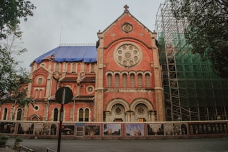 A large brick church with Romanesque architectural features, including circular rose windows and ornate stone detailing. The building is undergoing restoration, as indicated by the scaffolding and blue tarp visible on one side. Trees are present on both sides of the building, and there is a barrier displaying historic photographs in front of the church.