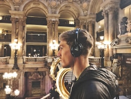 A person wearing headphones is standing in a grand, ornate hall with high columns and intricate architectural details. The space is warmly lit with classical chandeliers, creating a luxurious and historic atmosphere. Statues and decorative elements are visible, contributing to the opulent interior.