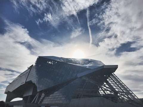 A modern, angular building with a reflective glass surface stands against a backdrop of a bright sky with scattered clouds. The sun casts a glow, creating interesting patterns in the clouds.