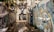 A group of pigeons is flying through an urban alleyway, lined with walls covered in colorful graffiti and street art. The alley is narrow with trees slightly overhanging. The graffiti features various designs and tags, adding a vibrant background to the scene.