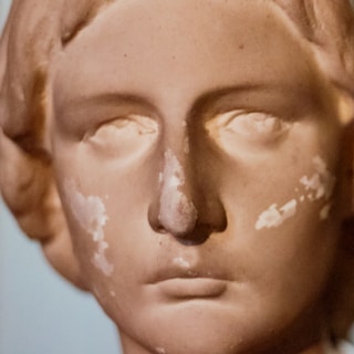 A close-up of a weathered classical sculpture featuring a serene face with closed eyes. The surface shows signs of wear and patches of discoloration.