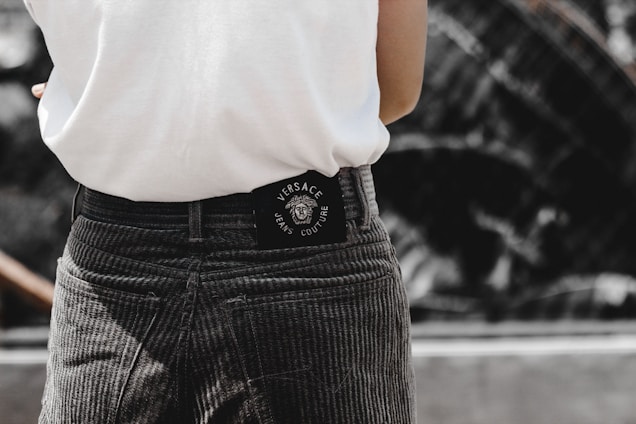 A person is wearing grey corduroy pants and a white shirt. The back of the pants has a black Versace Jeans Couture label. The background appears to be blurred, possibly an outdoor setting.