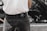 A person is wearing grey corduroy pants and a white shirt. The back of the pants has a black Versace Jeans Couture label. The background appears to be blurred, possibly an outdoor setting.