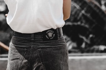 A person is wearing grey corduroy pants and a white shirt. The back of the pants has a black Versace Jeans Couture label. The background appears to be blurred, possibly an outdoor setting.