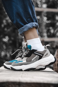 A pair of stylish sneakers is being worn with white socks and rolled-up blue jeans. The shoes feature a combination of grey, black, and teal colors with a prominent brand logo on the side. The setting appears to be outdoors, with a blurred natural background of greenery.