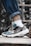 A pair of stylish sneakers is being worn with white socks and rolled-up blue jeans. The shoes feature a combination of grey, black, and teal colors with a prominent brand logo on the side. The setting appears to be outdoors, with a blurred natural background of greenery.