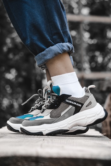 A pair of stylish sneakers is being worn with white socks and rolled-up blue jeans. The shoes feature a combination of grey, black, and teal colors with a prominent brand logo on the side. The setting appears to be outdoors, with a blurred natural background of greenery.