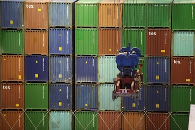 A large crane operates in front of a stacked wall of shipping containers in various colors such as blue, green, and brown. The containers are aligned in a grid pattern, and the crane apparatus is in the foreground, suggesting active loading or unloading operations in a shipping or industrial area.