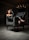 A woman is seated in a dark, elegant room on a luxurious upholstered chair with an intricate pattern. She is dressed in black leather attire and high-heeled shoes. There's a chandelier with lit candles in the background, adding a sophisticated touch to the ambiance.