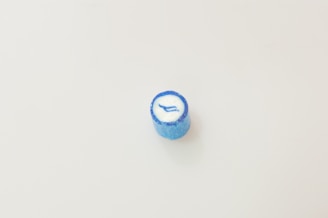 A single blue candy sits on a plain white background. The candy has a cylindrical shape with a glossy texture and a white center featuring a blue arrow-like design.