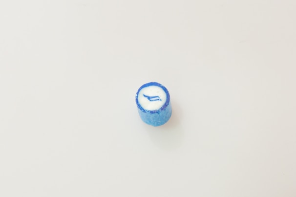 A single blue candy sits on a plain white background. The candy has a cylindrical shape with a glossy texture and a white center featuring a blue arrow-like design.