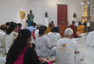 A group of people are seated on the floor in a room that appears to be a religious or cultural setting. Some are wearing traditional clothing while others are in casual attire. A few individuals, including monks dressed in orange robes, are facing an area displaying a golden shrine or statue. The walls of the room are white, and there are art pieces displayed around. A person standing appears to be taking photographs.