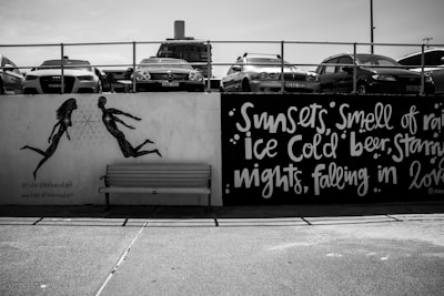 A black and white street art mural featuring silhouettes of a man and a woman running towards each other painted on a wall. Above them, cars are parked on a platform, and text on the wall reads phrases like 'Sunsets, Smell of rain, Ice Cold beer, Starry nights, falling in love.' There is a bench in front of the mural.