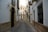 A narrow, winding cobblestone street flanked by traditional Spanish buildings featuring wrought iron balconies and ornate light fixtures. The architecture includes white walls with stone accents around doors and windowsills, while hanging signs indicate business establishments.