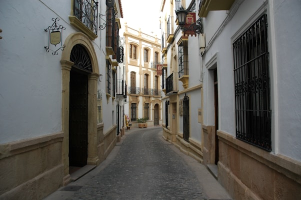 A narrow, winding cobblestone street flanked by traditional Spanish buildings featuring wrought iron balconies and ornate light fixtures. The architecture includes white walls with stone accents around doors and windowsills, while hanging signs indicate business establishments.