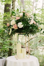 A large floral arrangement with a variety of white and pink flowers, surrounded by lush green leaves. The arrangement is perched atop a metallic gold geometric stand, which sits on a round white table. The background is a natural setting with green foliage and tree trunks visible.