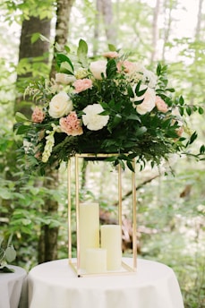 A large floral arrangement with a variety of white and pink flowers, surrounded by lush green leaves. The arrangement is perched atop a metallic gold geometric stand, which sits on a round white table. The background is a natural setting with green foliage and tree trunks visible.