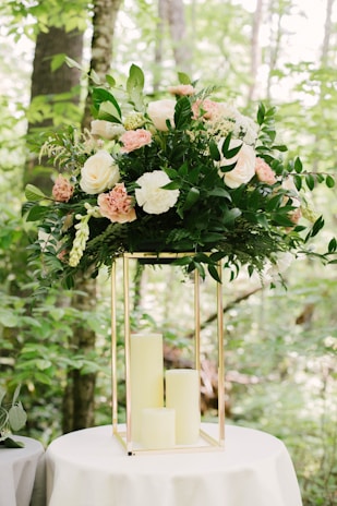 A large floral arrangement with a variety of white and pink flowers, surrounded by lush green leaves. The arrangement is perched atop a metallic gold geometric stand, which sits on a round white table. The background is a natural setting with green foliage and tree trunks visible.
