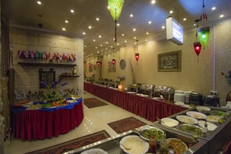 An indoor restaurant setting featuring a buffet arrangement with an assortment of dishes, plates, and cutlery. The room is decorated with small flags, red table skirts, and ornamental lighting. A table is set with various fruits and vegetables, and the wall is adorned with framed artwork.