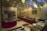 An indoor restaurant setting featuring a buffet arrangement with an assortment of dishes, plates, and cutlery. The room is decorated with small flags, red table skirts, and ornamental lighting. A table is set with various fruits and vegetables, and the wall is adorned with framed artwork.
