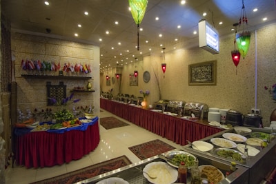 An indoor restaurant setting featuring a buffet arrangement with an assortment of dishes, plates, and cutlery. The room is decorated with small flags, red table skirts, and ornamental lighting. A table is set with various fruits and vegetables, and the wall is adorned with framed artwork.