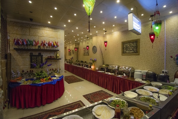 An indoor restaurant setting featuring a buffet arrangement with an assortment of dishes, plates, and cutlery. The room is decorated with small flags, red table skirts, and ornamental lighting. A table is set with various fruits and vegetables, and the wall is adorned with framed artwork.
