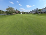 A beautifully maintained golf course with lush green grass and several people playing. To the right, there is a large white building with outdoor seating and several golf carts. On the left, there is a lake surrounded by trees and shrubs. The sky is clear with a few white clouds.