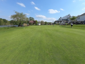 A beautifully maintained golf course with lush green grass and several people playing. To the right, there is a large white building with outdoor seating and several golf carts. On the left, there is a lake surrounded by trees and shrubs. The sky is clear with a few white clouds.