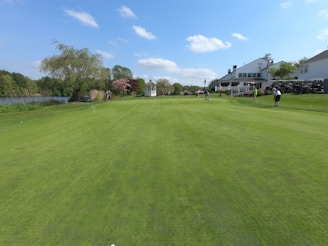 A beautifully maintained golf course with lush green grass and several people playing. To the right, there is a large white building with outdoor seating and several golf carts. On the left, there is a lake surrounded by trees and shrubs. The sky is clear with a few white clouds.