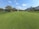 A beautifully maintained golf course with lush green grass and several people playing. To the right, there is a large white building with outdoor seating and several golf carts. On the left, there is a lake surrounded by trees and shrubs. The sky is clear with a few white clouds.