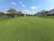 A beautifully maintained golf course with lush green grass and several people playing. To the right, there is a large white building with outdoor seating and several golf carts. On the left, there is a lake surrounded by trees and shrubs. The sky is clear with a few white clouds.