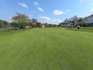 A beautifully maintained golf course with lush green grass and several people playing. To the right, there is a large white building with outdoor seating and several golf carts. On the left, there is a lake surrounded by trees and shrubs. The sky is clear with a few white clouds.