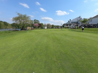 A beautifully maintained golf course with lush green grass and several people playing. To the right, there is a large white building with outdoor seating and several golf carts. On the left, there is a lake surrounded by trees and shrubs. The sky is clear with a few white clouds.