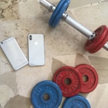 Two smartphones are placed on a tiled floor alongside a dumbbell with interchangeable weights. The dumbbell has blue and red weight plates, and the smartphones have sleek metallic finishes. The setting suggests a combination of technology and fitness.