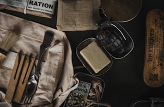 A collection of vintage military items including a ration pack, a black grenade, a bar of soap, a shaving brush, cutlery wrapped in cloth, matches, and a tin box. The items are arranged on a dark surface, suggesting a historical or military theme.