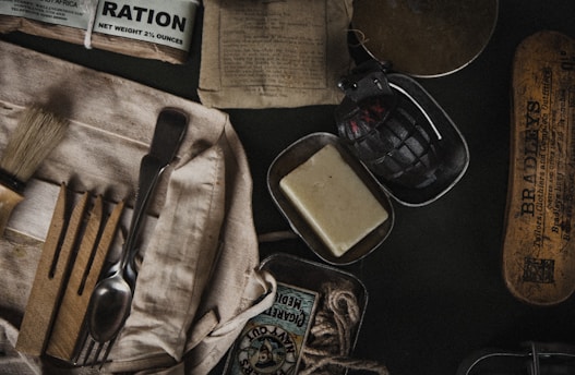 A collection of vintage military items including a ration pack, a black grenade, a bar of soap, a shaving brush, cutlery wrapped in cloth, matches, and a tin box. The items are arranged on a dark surface, suggesting a historical or military theme.