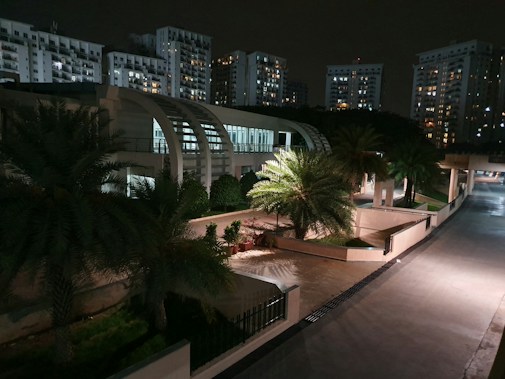 A nighttime urban scene with modern high-rise apartment buildings illuminated by their interior lights. In the foreground, there is a landscaped area with palm trees and a well-lit pathway. The architecture features smooth, curving lines and glass windows, creating a contemporary aesthetic.