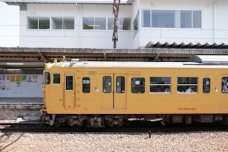 A yellow passenger train is stationed at a platform, with several people visible through the windows. Behind the train, there is a white building with large windows and posters displayed on the wall under a roofed area.