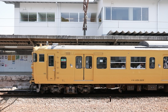 A yellow passenger train is stationed at a platform, with several people visible through the windows. Behind the train, there is a white building with large windows and posters displayed on the wall under a roofed area.