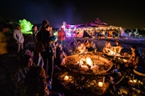 A lively outdoor gathering around a large campfire at night, with several people sitting close, possibly socializing. Candles are scattered around, adding to the warm glow of the scene. In the background, a tent, illuminated with colorful lights, hosts more people mingling under the night sky.