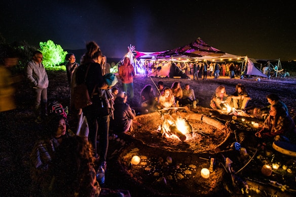 A lively outdoor gathering around a large campfire at night, with several people sitting close, possibly socializing. Candles are scattered around, adding to the warm glow of the scene. In the background, a tent, illuminated with colorful lights, hosts more people mingling under the night sky.