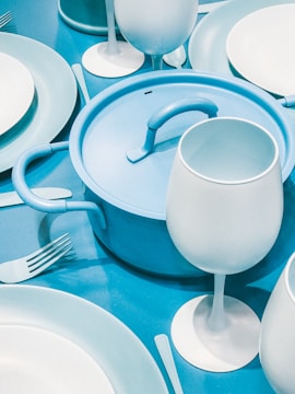 A neatly arranged tableware set in various shades of blue, featuring a pot with a handle, wine glasses, plates, forks, and knives. The color palette consists mostly of light blue and white, creating a harmonious and modern look.