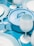 A neatly arranged tableware set in various shades of blue, featuring a pot with a handle, wine glasses, plates, forks, and knives. The color palette consists mostly of light blue and white, creating a harmonious and modern look.