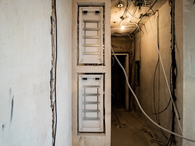 A partially constructed or undergoing renovation space featuring two white electrical panels on the left wall. The ceiling is exposed, showing various hanging wires and cables. The walls are unfinished, with visible patches and rough textures. The lighting is dim, primarily coming from a single bulb hanging from the ceiling.