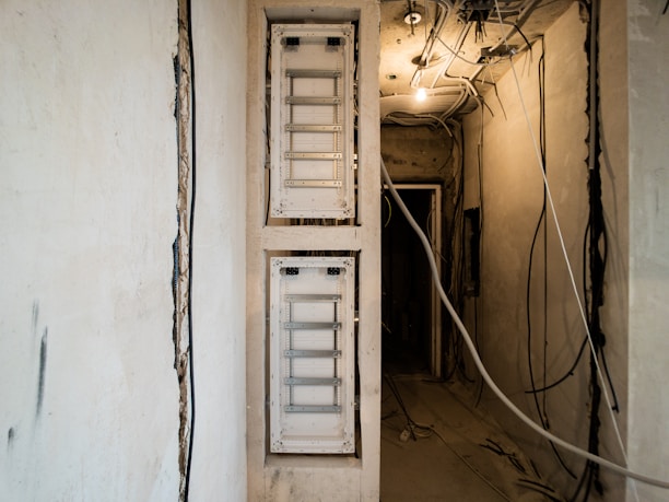 A partially constructed or undergoing renovation space featuring two white electrical panels on the left wall. The ceiling is exposed, showing various hanging wires and cables. The walls are unfinished, with visible patches and rough textures. The lighting is dim, primarily coming from a single bulb hanging from the ceiling.