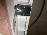 A metal bracket is attached to the edge of a textured surface. The bracket is secured by screws to a white plastic or composite material. A black cable is visible running in the background.