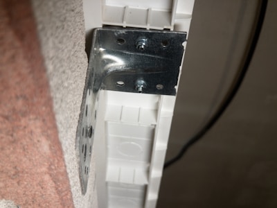 A metal bracket is attached to the edge of a textured surface. The bracket is secured by screws to a white plastic or composite material. A black cable is visible running in the background.
