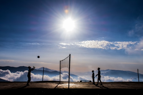 Silhouettes of people playing volleyball on a mountaintop, with a bright sun in the sky and a backdrop of clouds and distant mountains.
