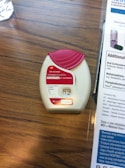 A medication inhaler labeled 'Anoro Ellipta' is placed on a wooden surface. The inhaler has a red and white design with clear dosage instructions and a counter displaying the number '12'. Beside it, part of a paper or booklet with informational text is visible.