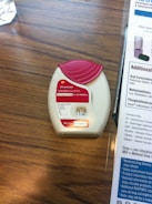 A medication inhaler labeled 'Anoro Ellipta' is placed on a wooden surface. The inhaler has a red and white design with clear dosage instructions and a counter displaying the number '12'. Beside it, part of a paper or booklet with informational text is visible.